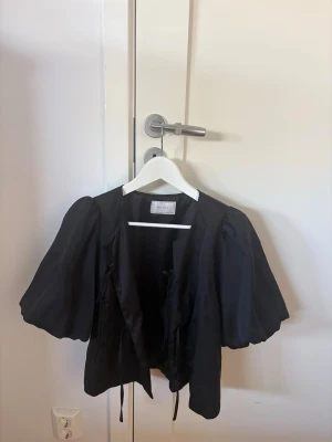 Svart blus med puffärmar från neo noir - Trendig svart blus från neo noir med stora puffärmar och knytband framtill. Blusen har en kortare, boxig passform och är tillverkad i ett glansigt tyg som ger en lyxig känsla. Perfekt för dig som vill sticka ut med en cool och modern look. Man kan använda som både tröja men även kofta