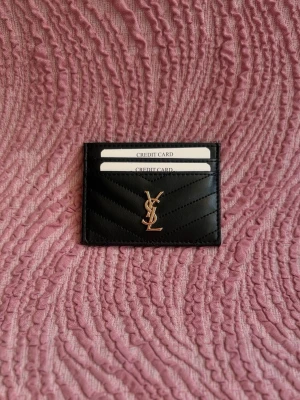 Svart YSL korthållare i läder - Stilren svart korthållare från YSL i äkta läder med guldigt logomärke framtill. Plats för flera kort och snyggt mönstrad yta. Perfekt accessoar för dig som vill ha något lyxigt och smidigt i fickan.