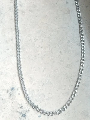 Kedjehalsband i silverfärg - Snyggt halsband med klassisk kedjedesign i silverfärgad metall. Länkarna är platta och tätt sammanlänkade vilket ger ett stilrent och modernt intryck. Perfekt accessoar för att lyfta din outfit.