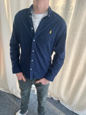 Mörkblå skjorta från Polo Ralph Lauren - Mörkblå skjorta från Polo Ralph Lauren i slim fit med klassisk krage och knappar framtill. Ikonisk gul broderad logga på bröstet. Personen på bilden är 183 cm lång. Skicka prisförslag 