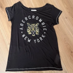 Svart Abercrombie t-shirt med tigertryck - Svart t-shirt från Abercrombie & Fitch med coolt tigertryck och texten 'Abercrombie New York' på framsidan. T-shirten har korta ärmar, rund hals och kontrastsömmar vid ärmslut och hals. Materialet är mjukt och lätt bomull. Har lite små hål. 