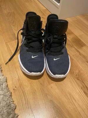 Svarta Nike sneakers med vit sula - Säljer ett par svarta Nike sneakers med vit sula och snygga detaljer. Skorna har snörning, ribbad mesh-ovandel och en stabil plattform. Swoosh-loggan syns tydligt på sidan och framtill. Perfekta för dig som gillar sportig och modern stil.