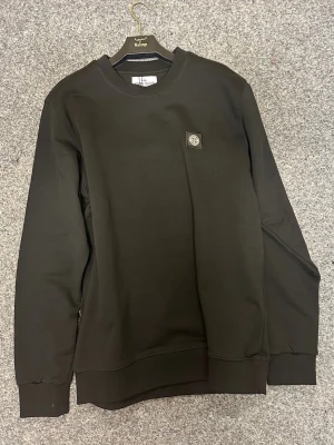 Svart sweatshirt från Stone Island - Svart crewneck sweatshirt från Stone Island med klassisk logopatch på bröstet. Tröjan har rund hals, långa ärmar och ribbade muddar vid ärmslut och nederkant. Tillverkad i mjuk bomull som är skön att bära och passar perfekt till jeans eller cargo.