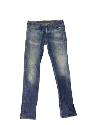 Blå Dsquared2 jeans - Säljer ett par blå jeans från Dsquared2 i storlek 34/34. Jeansen har slitna detaljer, lagad reva på knät och coola blekta partier. Klassisk femficksmodell med dragkedja och knapp. Perfekta för dig som gillar en avslappnad och trendig look.
