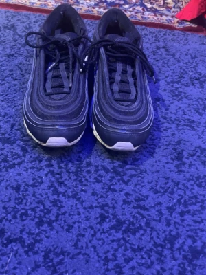 Svarta Nike Air Max 97 sneakers - Säljer ett par svarta Nike Air Max 97 sneakers med vita detaljer och synlig Air-sula. Skorna har en vågig design, snörning och rund tå. Ikonisk siluett med reflekterande inslag och slitstark syntet/mesh-överdel. Perfekta för dig som gillar streetwear och sportig stil.