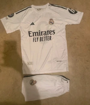 Real Madrid Mbappé matchställ - Säljer ett Real Madrid matchställ med Mbappé och nummer 9 på ryggen. Setet består av vit tröja och shorts med svarta detaljer och klubbens emblem. Materialet är lätt polyester som andas, perfekt för fotboll. Snygg design med tryckta loggor och sponsor på bröstet.