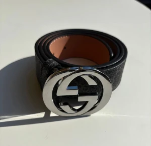 Svart Gucci bälte med GG-spänne - Snyggt svart bälte från Gucci med klassiskt GG-mönster och ett stort, rundat metallspänne i silver. Bältet är gjort av skinn med en mörkgrå/svart yta och har en lyxig känsla. Insidan är ljusbrun och har präglad Gucci-text. Perfekt för att lyfta din outfit.