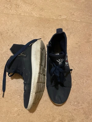 Mörkblå sneakers med dragkedja - Säljer ett par mörkblå sneakers med vita sulor och snörning. Skorna har rund tå och dragkedja på sidan för extra stil och enkel påtagning. Materialet är syntet och de har en platt sula. Perfekt för dig som gillar sportig och avslappnad look.