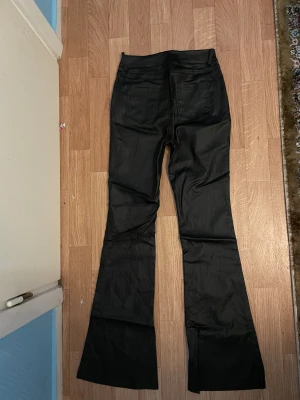 Svarta bootcut byxor med hög midja läder  - Snygga svarta byxor med bootcut-ben och hög midja. De har klassiska fickor fram och bak samt bälteshällor. Materialet har en lätt glansig finish som ger en cool vibe. Perfekta för dig som gillar en trendig siluett med lite extra attityd.
