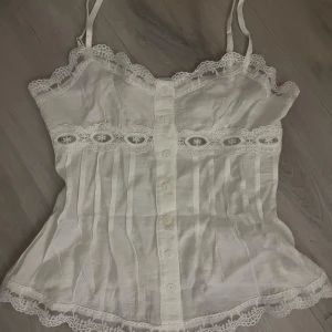 Vitt spetslinne 🤞 - Charmig vit linnetopp med spetsdetaljer och knappar framtill. Toppen har smala axelband och dekorativa spetskanter längs halsringning.