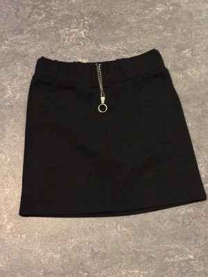 Svart kortkjol med dragkedja - Stilren svart kortkjol med markerad midja och en snygg silverfärgad dragkedja framtill med ringdetalj. Kjolen har en rak passform och är perfekt för dig som gillar minimalistisk stil. Materialet känns mjukt och följsamt.