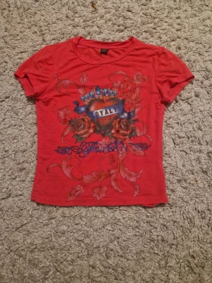 Rosa t-shirt med tryck från SHEIN - Cool rosa t-shirt från SHEIN i storlek XS med ett stort tryck framtill som visar ett hjärta, rosor och texten 'LOYALTY' under en krona. T-shirten har korta ärmar och är gjord i mjuk bomull. Passformen är normal och trycket har detaljer i blått och rött. Ser röd ut i kameran men är skrikrosa