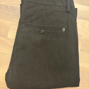 Mörkbruna chinos från Dondup - Mörkbruna chinos från Dondup med klassisk passform och raka ben. Diskreta bakfickor med knapp samt litet D-emblem. Tillverkade i bomull med en lätt stretch – jättesköna att ha på sig. Mått: midja 43 cm, längd 95 cm, benslut 15 cm, insida ben 24 cm.