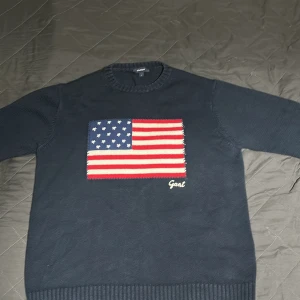 Mörkblå stickad tröja från Gant - Snygg mörkblå stickad tröja från Gant med amerikansk flagga broderad på bröstet och Gant-logga i vitt. Tröjan har rund hals och långa ärmar med ribbade muddar. Perfekt för dig som gillar klassisk och cool stil.