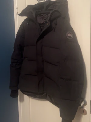 SÖKER BYTEN MOT JACKA! - Svart pufferjacka från Canada Goose med huva och tydlig logotyp på ärmen. Jackan har grova dragkedjor, ribbade muddar och quiltad design. Perfekt för kalla dagar med sin tjocka dunfyllning och stilrena look. Snygg passform och klassisk vinterstil.