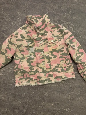 Camouflage jeansjacka med fransad kant - Säljer en jeansjacka med rosa, grön och beige camouflage-mönster. Jackan har knappar framtill, två bröstfickor med lock och en fransad nederkant som ger en cool vibe. Perfekt för dig som vill sticka ut med din stil.