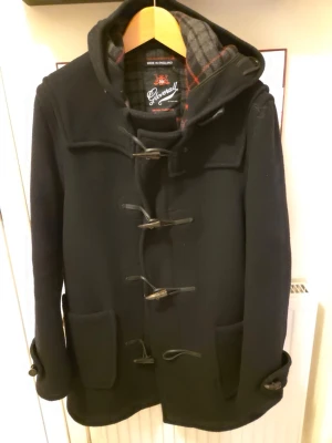 Svart duffel från Gloverall, stl 40 - Klassisk svart duffeljacka från Gloverall med huva och rutigt innerfoder i grått och rött. Jackan har träknappar med snörning framtill, stora fickor och är tillverkad i ull. Perfekt för kyliga dagar och tidlös brittisk stil. 