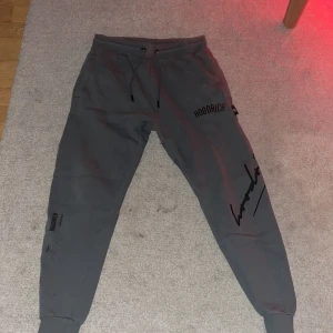 Grå mjukisbyxor från Hoodrich - Snygga grå mjukisbyxor från Hoodrich med svart och röd logga. Byxorna har snörning i midjan och muddar vid bensluten för en bekväm passform. Perfekta för en avslappnad stil.