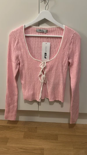 Rosa ribbad kofta från FB Sister - Supersöt ljusrosa kofta från FB Sister med ribbad struktur och små hålmönster. Tröjan har lång ärm, croppad passform och knyt framtill med vit snörning. Perfekt för dig som gillar gulliga och trendiga plagg med lite extra detalj.