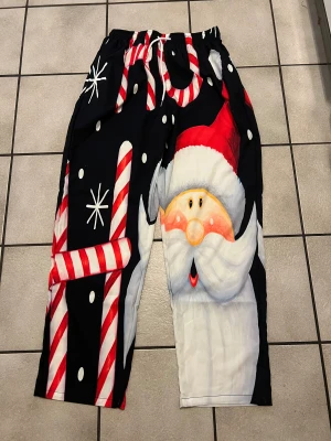 Julbyxor med tomtemotiv och polkagrisar - Säljer ett par svarta byxor i polyester med färgglatt tryck av tomte och polkagrisar. Byxorna har snörning i midjan och vita detaljer som snöflingor. Perfekt för dig som vill sticka ut på julfirandet!