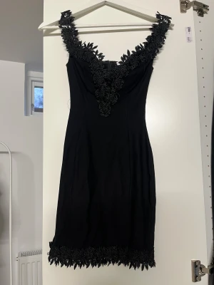 Svart kort klänning med spetsdetaljer - Supersnygg svart klänning med v-ringad hals och blommiga spetsdetaljer längs urringningen och nederkanten. Klänningen är ärmlös och har en figurnära passform som framhäver siluetten. Perfekt för dig som vill sticka ut med eleganta detaljer.