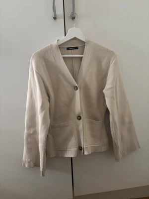 Beige kofta från Gina Tricot - Snygg beige kofta från Gina Tricot i mjuk bomull. Modellen har v-ringning, tre stora knappar framtill och två praktiska fickor. Ärmarna är raka och lite bredare, och koftan har ribbade detaljer vid muddar och nederkant. Perfekt för lager-på-lager under kyliga dagar. Helt ny, råkade ta av prislappen så kan inte lämna tillbaka. 