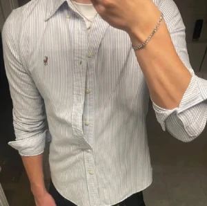 Randig skjorta från Polo Ralph Lauren - Säljer en klassisk skjorta från Polo Ralph Lauren i vit och blå smalrandig design. Skjortan har lång ärm, knappar framtill och den ikoniska broderade loggan på bröstet. Tillverkad i mjuk bomull och har en normal passform. Perfekt för dig som gillar stilrena och tidlösa plagg. ⭐️ny pris 1700kr