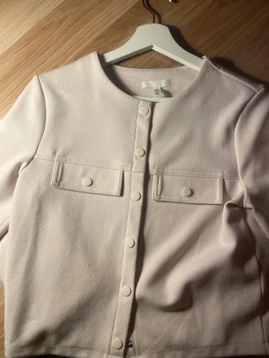 Ljusbeige overshirt från H&M - Snygg ljusbeige overshirt från H&M med rund hals och tryckknappar framtill. Jackan har två bröstfickor med lock och är tillverkad i mjuk bomullsmix. Perfekt att slänga över en t-shirt för en clean look.