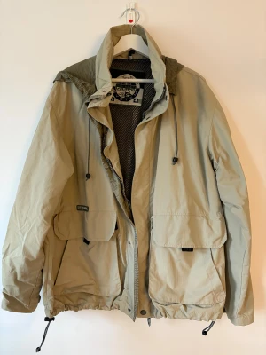 Beige vindjacka Colmar med huva - Snygg beige vindjacka från Colmar med meshfoder, justerbar huva och stora fickor framtill. Jackan har dragsko i midjan och vid ärmslut, samt dragkedja och tryckknappar. Perfekt för dig som gillar sportig och avslappnad stil.Oversized