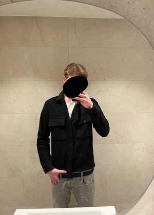 Svart overshirt med fickor - Säljer en svart overshirt med klassisk krage och två stora bröstfickor med lock och knapp. Jackan har knäppning framtill och raka ärmar. Perfekt att slänga över en t-shirt för en clean och enkel look. Modellen på bilden är 186 och väger 72 kg och den sitter perfekt på han.