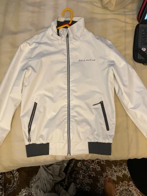 Vit vindjacka från Race Marine - Snygg vit vindjacka från Race Marine med svart dragkedja och svarta detaljer vid fickorna. Jackan har hög krage, långa ärmar och elastiska muddar i svart vid nederkant och ärmslut. Perfekt för dig som gillar stilren och sportig look.