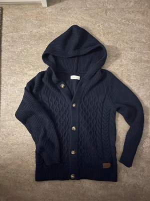 Marinblå stickad kofta med huva Pier One - Snygg marinblå stickad kofta från Pier One med huva och grova kabelstickade detaljer framtill. Koftan har långa ärmar, ribbade muddar och knäppning med fem knappar. Perfekt för dig som gillar chill och stilren look.