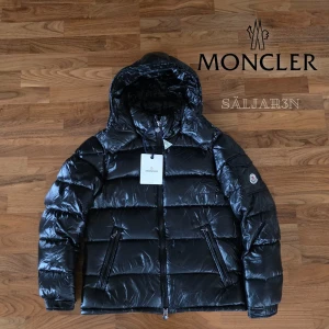Moncler Maya Jacka - Exklusiv Moncler Maya-jacka i svart, storlek M. Nyskick med taggar! Ikonisk modell med blank finish och quiltad design. Fylld med premium-dun som ger värme och komfort utan att kännas tung. Utrustad med fast huva, dragkedjeförsedda fickor samt klassiskt Moncler-märke på ärmen. Insidan pryds av den ikoniska Moncler-seriekomikern. En tidlös och lyxig jacka som kombinerar stil och funktion – perfekt för vintern. 📦 Snabb leverans – skickas samma dag!