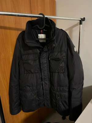 Svart Moncler pufferjacka herr - Svart pufferjacka från Moncler med hög krage, fyra framfickor med lock och knappar, samt klassisk Moncler-logga på ärmen. Jackan har quiltade detaljer och dragkedja framtill. Materialet är syntet och jackan är perfekt för kyliga dagar.  Size 5 fits M-L.  100% Real