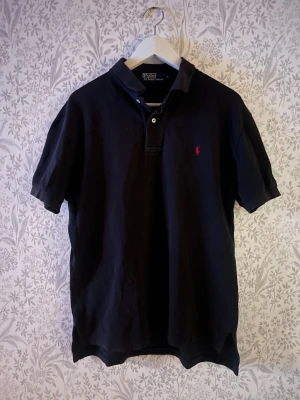 Ralph Lauren piké - Klassisk svart pikétröja från Polo Ralph Lauren. Tröjan har korta ärmar, krage och knappar framtill. På bröstet finns den ikoniska röda Polo-loggan. Notera att tyget är något solblekt. Axelbredd 48cm, längd 67cm. JAG SKICKAR INGA BILDER NÄR JAG BÄR PLAGGET:) OBS se defekt som är nedanför kragen baktill på sista bild.  !!