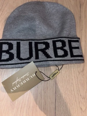 Grå mössa från Burberry - Snygg grå mössa från Burberry med stor svart logotyp längs kanten. Stickad i mjukt material och har en klassisk form som passar perfekt till kalla dagar. Mössan har en neutral färg som är lätt att matcha med olika outfits.