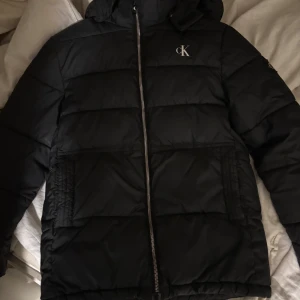 Svart pufferjacka Calvin Klein Jeans - Svart pufferjacka från Calvin Klein Jeans med huva och dragkedja framtill. Jackan har stora fickor, diskret CK-logga på bröstet och en patch på ärmen. Perfekt för kalla dagar och riktigt snygg streetstil. Jackan är knappt använd. Storlek M. Ge prisförslag 