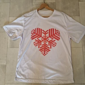 Vit t-shirt med rött hjärtmönster - Vit t-shirt med ett stort rött grafiskt hjärtmönster både fram och bak. T-shirten har klassisk rund hals och korta ärmar. Materialet är polyeste, perfekt för en chill och stilren look.