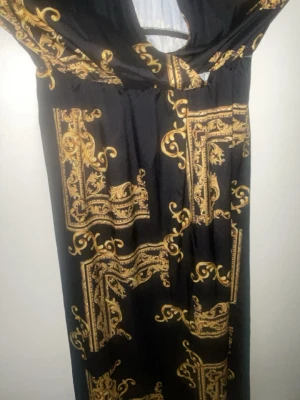 Svart långklänning med guldmönster - Säljer en svart långklänning med lyxigt barockinspirerat mönster i guld. Klänningen har en djup v-ringning och är ärmlös, vilket ger en elegant och trendig look. Perfekt för dig som vill sticka ut med stilrena detaljer och cool design.