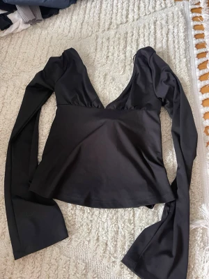 Svart omlottopp med v-ringning - Säljer en svart omlottopp med djup v-ringning och långa ärmar. Toppen har ett brett knytband i midjan som kan knytas framtill för en snygg siluett. Materialet är mjukt och har en lätt stretch, perfekt för att skapa en cool look.