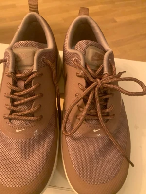 Nike Air Max Thea sneakers i brunt - Säljer ett par Nike Air Max Thea sneakers i en snygg brun färg med vita sulor. De är nya . Skorna har mesh- och syntetmaterial, klassisk Nike-logga på sidan och snörning framtill. Perfekta för dig som gillar stilrena och bekväma sneakers med sportig vibe.