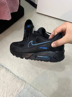 Nike Air Max 90 svart/blå sneakers BYTE! - Säljer ett par svarta Nike Air Max 90 sneakers med blå detaljer och swoosh. Skorna har en klassisk siluett, synlig Air Max-dämpning i sulan och är tillverkade i en mix av mesh och syntetmaterial. Perfekta för dig som gillar streetwear och sportig stil.