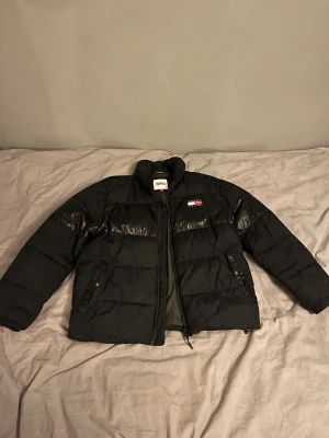 Svart pufferjacka Tommy Jeans M - Svart pufferjacka från Tommy Jeans i storlek M. Jackan har en hög krage, två fickor med dragkedja och quiltade paneler. Materialet är polyester och insidan är olivgrön. Klassisk Tommy Jeans-logga på bröstet. Perfekt för kalla dagar. Priset kan diskuteras vid smidig affär!