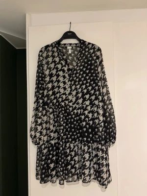 Svartvit mönstrad klänning - Skir klänning i svartvitt mönster. Svart tillhörande underklänning. Knappar ända upp i halsen och knappar i ärmslut.