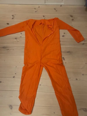 Orange jumpsuit med tryck - Säljer en helorange jumpsuit med långa ärmar och ben, dragkedja framtill och svart tryck med siffrorna 696969 på bröstet. Materialet känns lätt och syntetiskt, perfekt för dig som vill sticka ut med en färgstark look. Denna är perfekt för halloween om man vill klä ut sig till fånge. Passar mest för S men även för M, vilket jag är. 