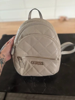 Beige quiltad ryggsäck från Guess - Snygg beige ryggsäck från Guess med quiltad design och logomönster över hela väskan. Väskan har två fack med dragkedja, justerbara axelband och metallogo framtill. Tillverkad i glansigt syntetmaterial som ger en lyxig känsla. Finns några små fläckar på sidan som jag ej vet om de går bort men inget som stör eller syns vidare mycket :) finns lite slitage uppe vid kanten