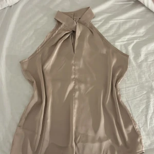 Beige omlott blus från SHEIN - Säljer en beige omlott blus från SHEIN i storlek S. Blusen är ärmlös med hög krage och två dekorativa knappar i halsen. Materialet är glansigt och känns som satin, vilket ger en lyxig känsla. Perfekt för dig som gillar stilrena och eleganta plagg.