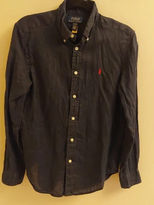 Mörkblå linneskjorta Polo Ralph Lauren ungdom - Klassisk mörkblå skjorta från Polo Ralph Lauren i linne med knappar framtill och ikonisk röd broderad logga på bröstet. Skjortan har lång ärm och button-down krage, perfekt för en clean och stilren look. Storlek 14-16 passar en 13-14 åring