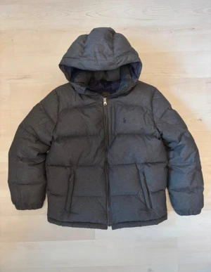 Mörkgrå dunjacka från Polo Ralph Lauren - Säljer en mörkgrå dunjacka från Polo Ralph Lauren med huva och dragkedja framtill. Jackan har broderad logga på bröstet, två sidofickor med dragkedja och en bröstficka. Puffad design och elastiska muddar vid ärmslut. Perfekt för kalla dagar!