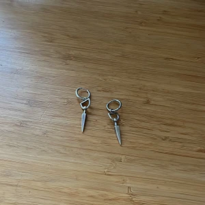 Silvriga Maria Nilsdotter örhängen med spetsig hängande detalj - Säljer ett par coola silvriga örhängen med rund ring och hängande spets. Jätte snygga och passar till allt! Ens örhänget har en lite mörkare ring men det ser man knappt.